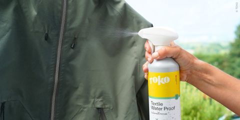 Im Freien sprüht eine Hand „Toko Textile Water Proof“ auf eine grüne Jacke.