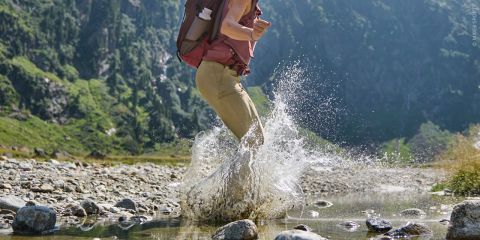 Eine Person mit einem Rucksack watet durch einen seichten Bach, umgeben von Felsen und Grün in einer bergigen Landschaft.