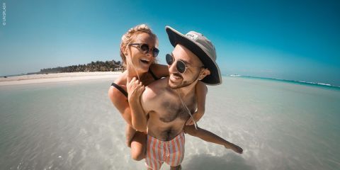 Eine Frau trägt einen Mann auf dem Rücken an einem sonnigen Strand; beide lächeln und tragen Sonnenbrillen, im Hintergrund sieht man das klare blaue Wasser und den blauen Himmel.