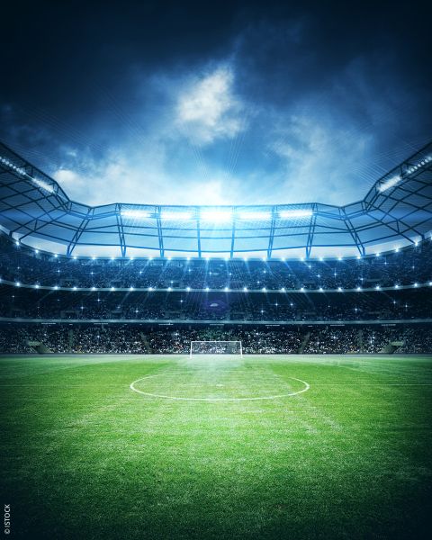 Dramatischer Blick auf ein beleuchtetes Fußballstadion mit grünem Spielfeld und Torpfosten, gefüllt mit jubelnden Fans unter einem bewölkten Abendhimmel.