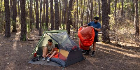 Zwei Menschen, die im Wald zelten; einer sitzt im Zelt, während der andere draußen steht und einen orangefarbenen Schlafsack in der Hand hält.