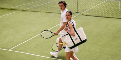 Ein Mann und eine Frau in weißen Tennisoutfits gehen über einen Rasenplatz, halten Schläger und eine große Tragetasche in der Hand und blicken zurück in die Kamera.