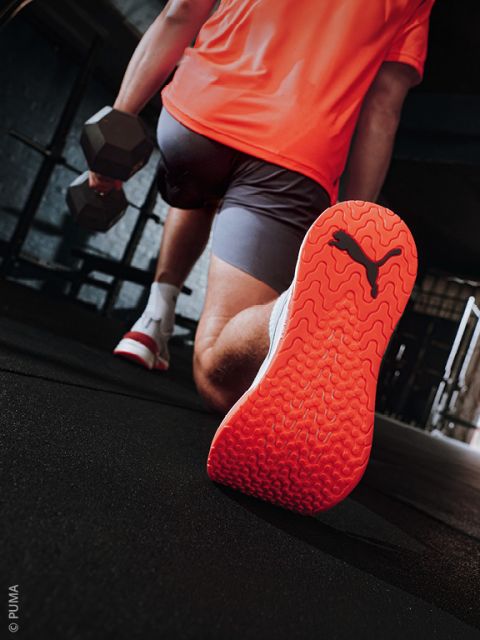 Eine Person macht drinnen eine Ausfallschrittbewegung, hält dabei eine Hantel in der Hand, trägt ein leuchtend orangefarbenes Oberteil und Puma-Sneaker mit roter Sohle und blickt in die Kamera.