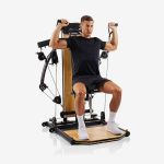 Ein Mann, der auf einem multifunktionalen Heimfitnessgerät trainiert, im Sitzen die Griffe über seine Schultern hebt und schwarze Trainingskleidung sowie weiße Socken trägt.
