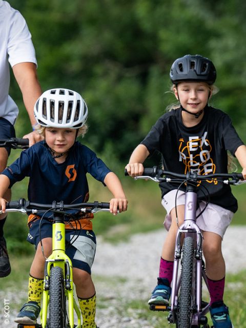 Zwei kleine Kinder mit Helmen fahren draußen auf einem Schotterweg Fahrrad, während ein Erwachsener in der Nähe steht. Im unscharfen Hintergrund ist Grün zu sehen.