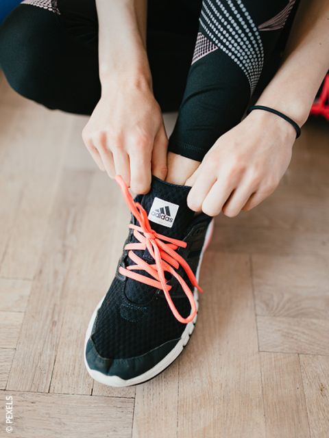 Eine Person, die leuchtend pinkfarbene Schnürsenkel an einem schwarzen Adidas-Sneaker bindet und dabei schwarze Leggings mit weißen und pinkfarbenen Mustern trägt.