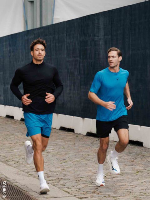Zwei Männer in Sportkleidung joggen auf einem Kopfsteinpflasterweg neben einem dunklen Zaun; beide tragen blaue Shorts und weiße Turnschuhe.
