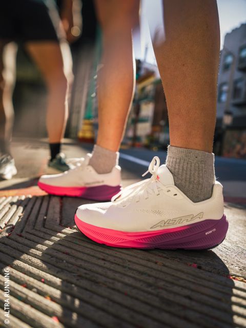 Eine Person in weißen und rosa Laufschuhen steht auf einem Bürgersteig in der Stadt; das Sonnenlicht lässt die Schuhe und Beine hervortreten, während der städtische Hintergrund verschwommen wirkt.
