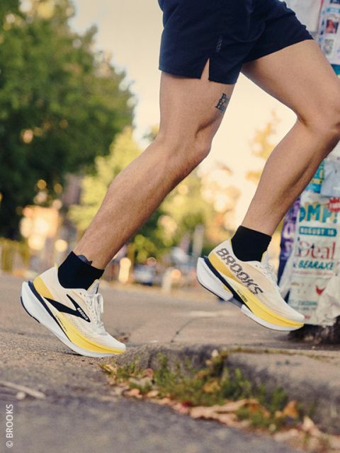 Ein Läufer in Shorts und gelb-weißen Brooks-Laufschuhen steigt auf einen Bordstein. Im Hintergrund sind Bäume und ein mit Plakaten beklebter Pfosten zu sehen.