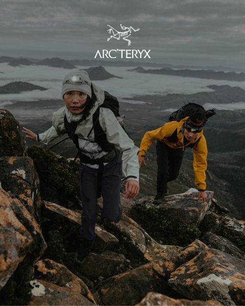 Zwei Personen wandern auf einem felsigen Bergpfad. Der Himmel ist bedeckt, im Hintergrund ist eine neblige Bergkette zu sehen. Beide Wanderer tragen Outdoor-Ausrüstung. Rechts befindet sich ein kleiner Teich. Oben ist das Arc'teryx-Logo zu sehen.