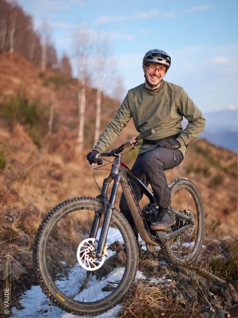 Eine Person mit Helm und grüner Jacke lächelt, während sie mit einem Mountainbike auf einem felsigen, verschneiten Weg vor dem Hintergrund von Bäumen posiert.