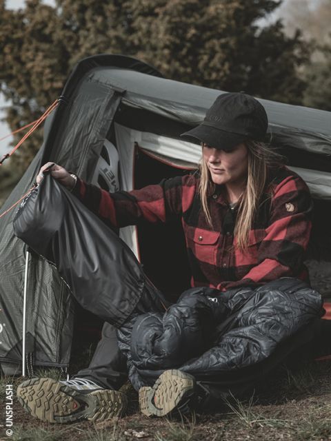 Eine Frau in einem roten Karohemd sitzt vor einem Zelt, zieht sich eine Regenhose und Wanderstiefel an und bereitet sich auf Aktivitäten im Freien vor.