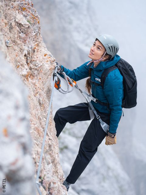 Eine Frau in Kletterausrüstung klettert an einem Sicherheitsseil gesichert eine felsige Klippe hinauf und blickt konzentriert nach oben. Im Hintergrund liegt Nebel.