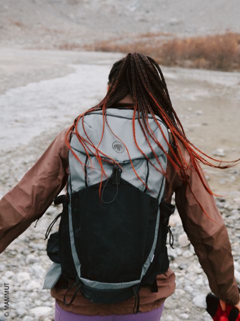 Eine Person mit geflochtenen Haaren und einem großen Rucksack läuft auf felsigem Gelände in der Nähe eines Flusses, den Rücken zur Kamera gewandt.