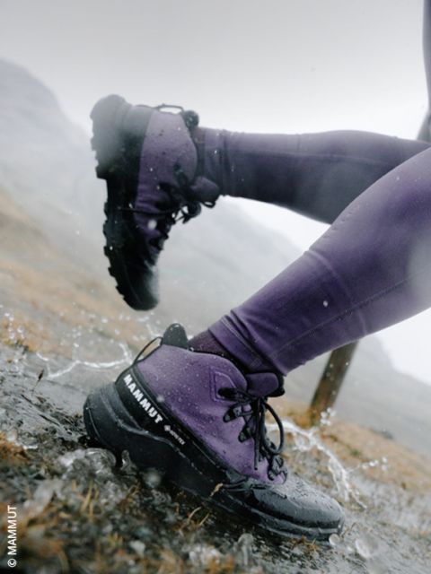 Nahaufnahme der Beine einer Person in einer lila Hose und schwarzen Wanderstiefeln, die auf einem regennassen, felsigen Wanderweg durch eine Pfütze watet.