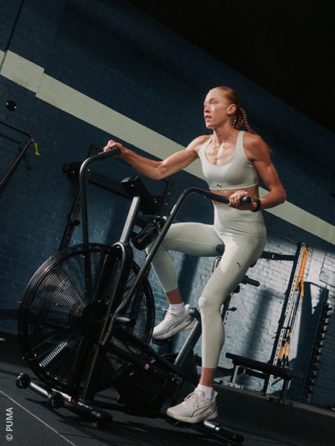 Ein Mann, der eine geblümte Kappe, ein beiges T-Shirt und rosa Shorts trägt, lächelt, während er auf einem stationären Heimtrainer der Marke Schwinn fährt.