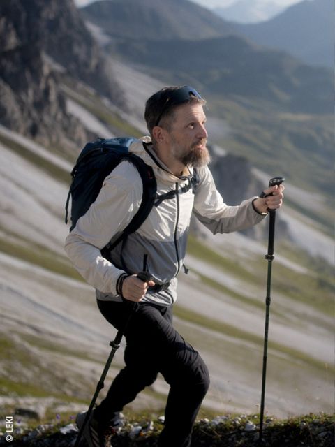 Ein Mann mit Bart wandert auf einem felsigen Bergpfad bergauf, wobei er Wanderstöcke benutzt und einen Rucksack trägt; im Hintergrund sind Berge zu sehen.