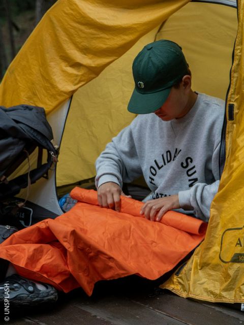 Eine Person mit einer grünen Mütze sitzt in einem gelben Zelt und faltet eine orangefarbene Isomatte zusammen, während sie im Freien zeltet.