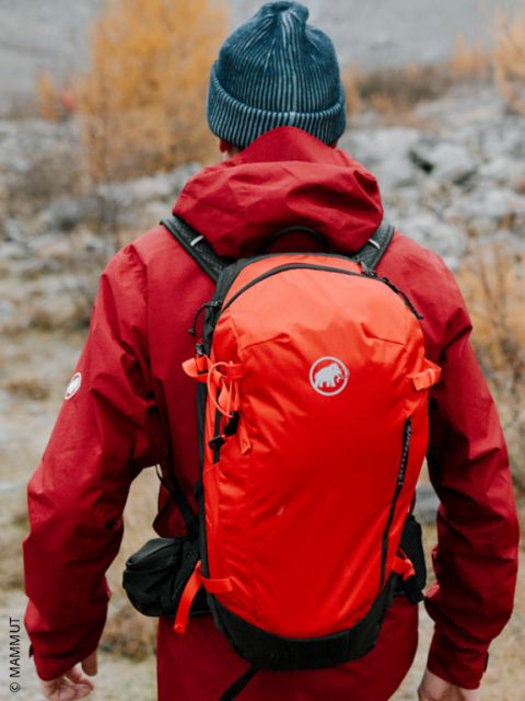 Eine Person in einer roten Jacke und einer blauen Mütze, die mit einem orangefarbenen Rucksack in der Natur wandert, von hinten gesehen.