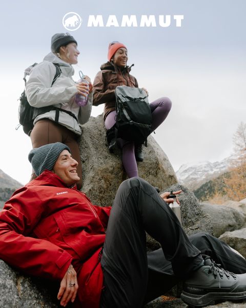 Zwei Wanderer in Outdoor-Ausrüstung durchqueren felsiges Gelände unter einem bewölkten Himmel, im Hintergrund ein großer Berg. Beide tragen Mammut-Kleidung, und das Markenlogo ist oben im Bild zu sehen.