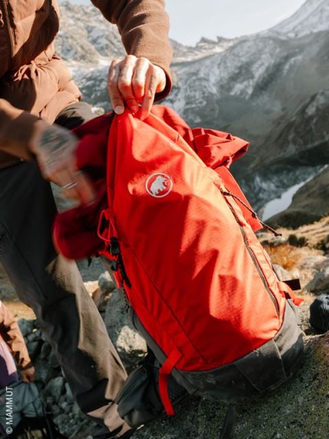 Eine Person in Winterkleidung und gemusterten Handschuhen legt eine Schneeschaufel in einen rosafarbenen Rucksack auf schneebedecktem Boden, mit verschneiten Bäumen im Hintergrund.