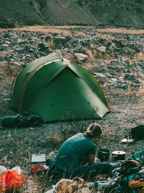 Eine Person in roter Jacke und Mütze bereitet im Freien Essen zu, im Gras hockend, in der Nähe von Campingausrüstung, gesehen durch die offene Tür eines Zeltes mit Bergen und Bäumen im Hintergrund.