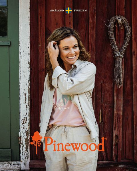 Ein Mann und eine Frau in Outdoor-Kleidung gehen unter einem bewölkten Himmel durch eine Wiese. Die Frau trägt eine schwarze Tasche, während der Mann einen Hut hält. Über ihnen sind das Pinewood-Logo und der Text „Scandinavian Outdoor Life“ zu sehen.