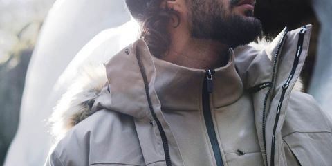 Mann in brauner Jacke, schwarzer Hose und grauer Mütze wandert durch einen dichten Wald. Er trägt schwarze Handschuhe und einen Rucksack. Der Wald hat hohe Bäume und abgefallene Blätter. „Rukka“-Logo oben.