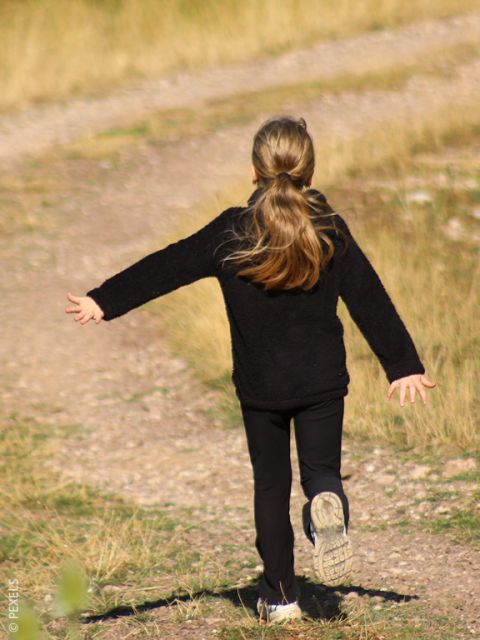 Ein Mädchen mit langen Haaren in einem schwarzen Outfit rennt fröhlich mit ausgestreckten Armen einen von Gras gesäumten Feldweg entlang.