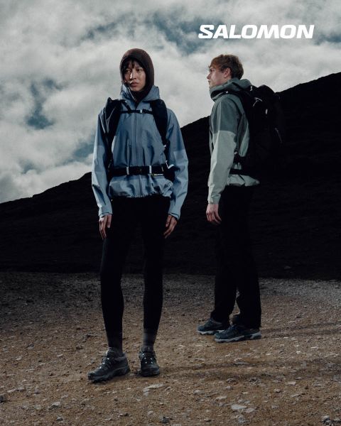 Nahaufnahme einer Person in Salomon-Schuhen und Salomon-Socken mit Farbverlauf in Blau. Sie steht auf einer Wiese mit einer malerischen Berglandschaft im Hintergrund. Ein Wanderer in Outdoor-Kleidung steht weiter entfernt, ebenfalls auf der Wiese.