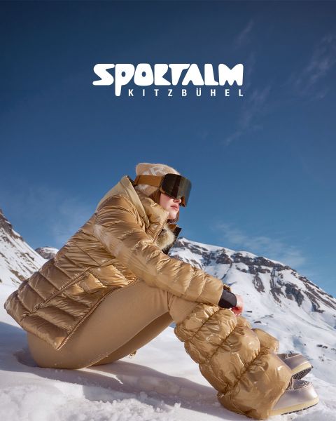 Eine Person in einer metallischen Pufferjacke und schwarzen Hosen liegt auf schneebedecktem Gelände mit Bergen im Hintergrund unter einem bewölkten Himmel. Die Szene ist ruhig und winterlich. In der oberen rechten Ecke ist der Text „SPORTALM KITZBÜHEL“ zu sehen.