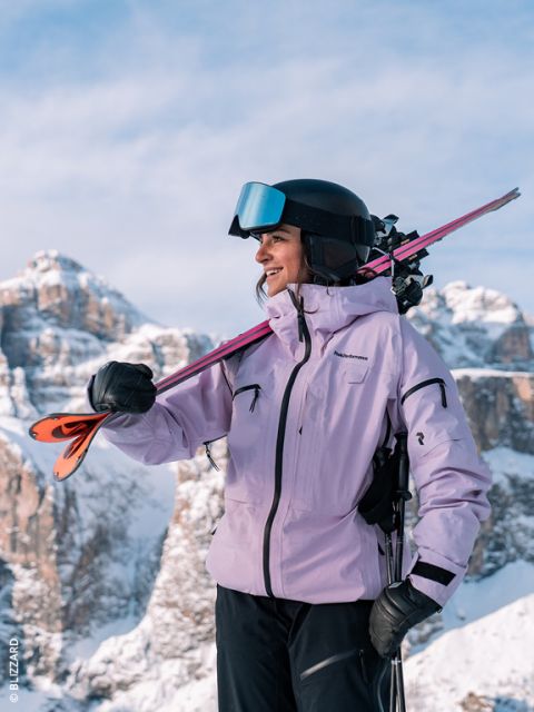 Eine Skifahrerin in einer lavendelfarbenen Jacke und mit Helm lächelt, während sie ihre Skier auf der Schulter trägt; im Hintergrund sind schneebedeckte Berge zu sehen.