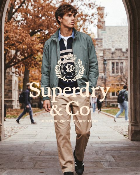 Ein Mann in einer grünen Jacke und einer khakifarbenen Hose geht auf einem Weg auf dem Campus; das Bild ist mit dem Logo „Superdry & Co“ überlagert.