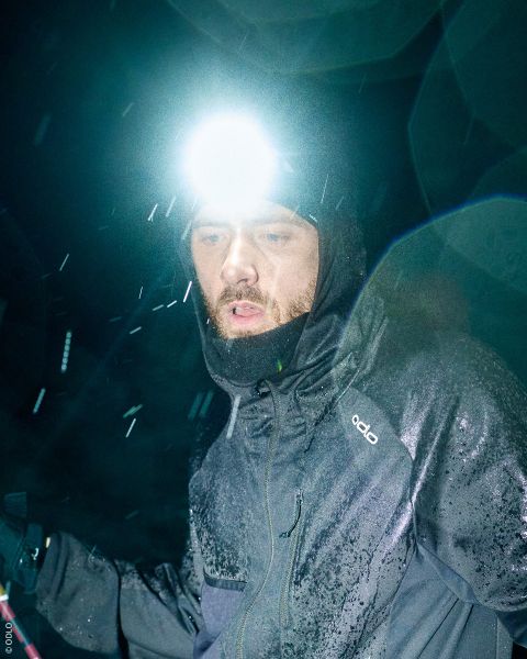 Eine Frau, die eine Stirnlampe, ein gemustertes Stirnband und eine Trailrunning-Weste trägt, blickt gegen den bewölkten Himmel empor.