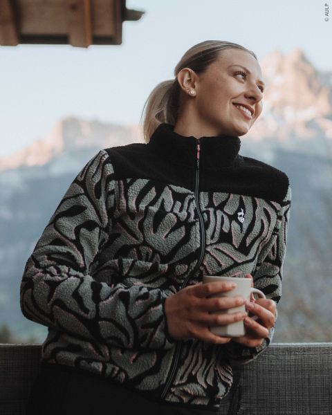 Eine Frau, die eine gemusterte Fleecejacke mit Reißverschluss trägt, hält eine Tasse in der Hand und blickt auf die Berge im Hintergrund.